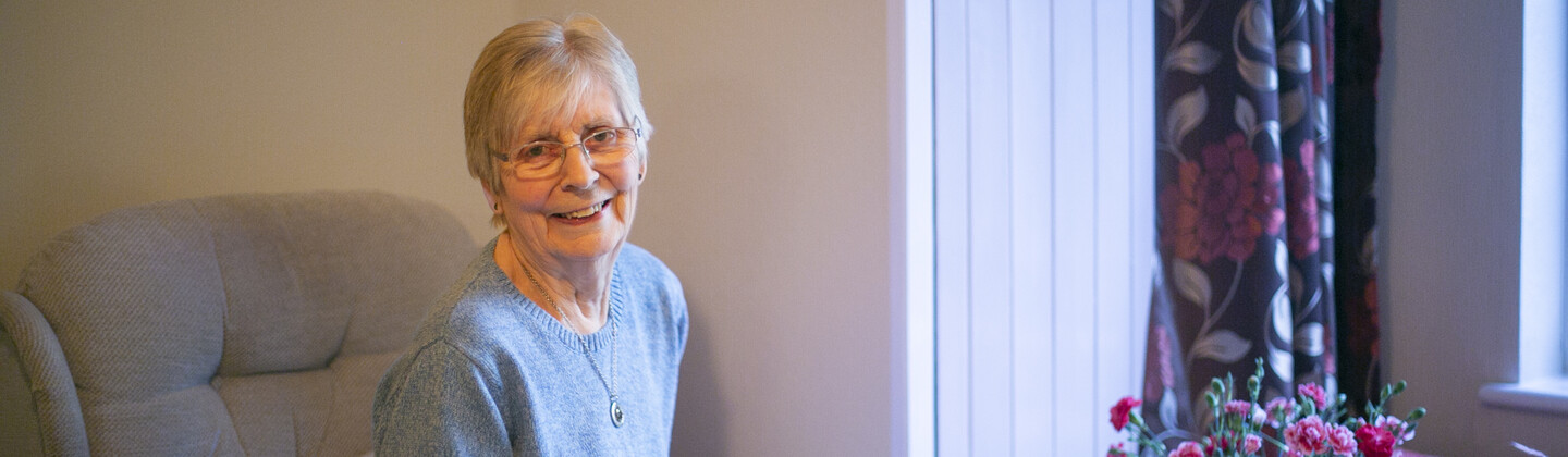 Woman is sitting in an arm chair writing in a book. She has a table in front of her with flowers in a vase.