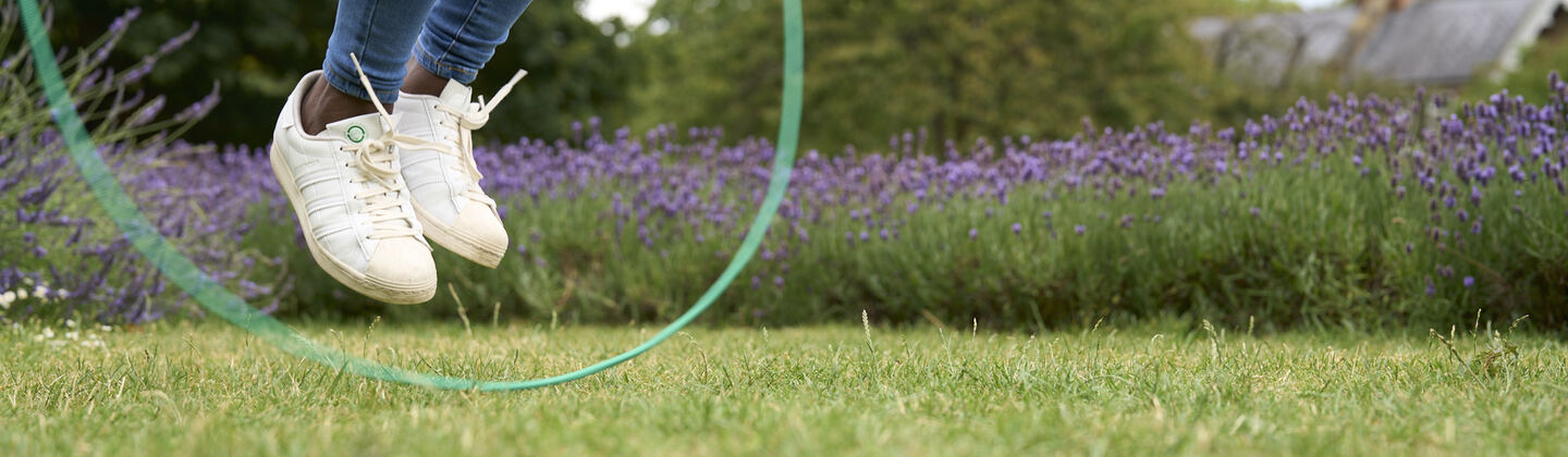A person is skipping rope in a grassy field. They are captured mid jump, with the rope just under their feet. Behind them is a natural fence of lavender plants.