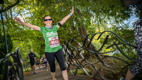 A woman jogging across a small bridge with her arms in the air, wearing a green Macmillan running t-shirt