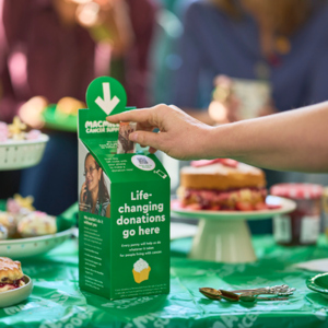 A person is donating £10 in a Coffee Morning donation box. It is on a table full of delicious bites and treats. 