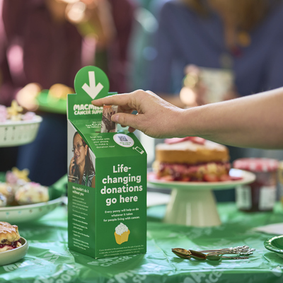 A person is donating £10 in a Coffee Morning donation box. It is on a table full of delicious bites and treats. 