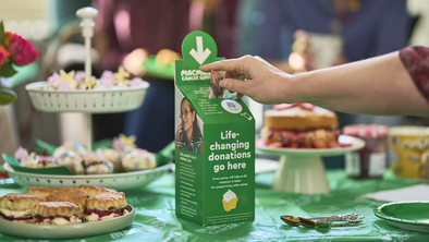 A person is donating £10 in a Coffee Morning donation box. It is on a table full of delicious bites and treats. 