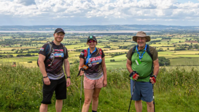 Three people are standing together in a field. Behind them is a view of the valley below.
