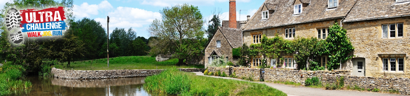 Countryside with a stone house covered in greenery, a small stone bridge over a calm stream, and trees under a blue sky with clouds.