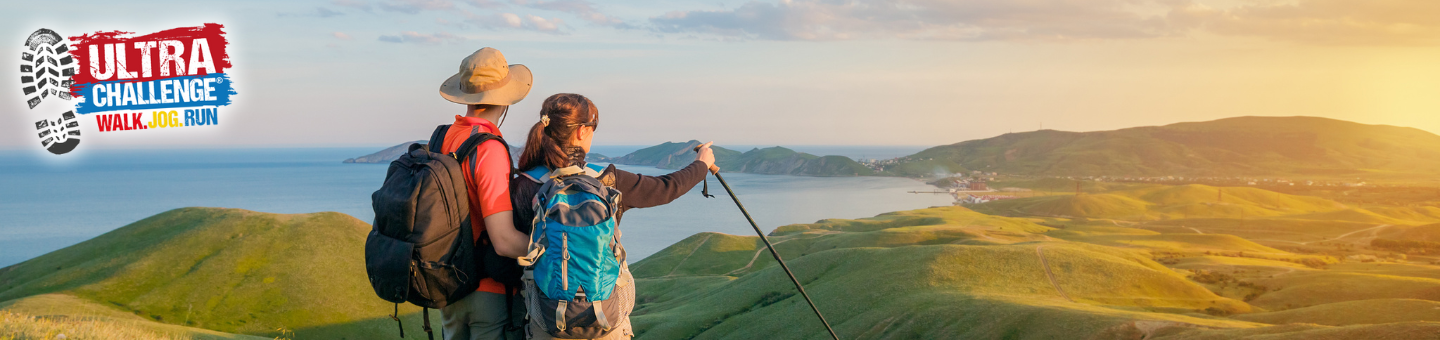 Two hikers with backpacks stand on a grassy hill at sunset, overlooking rolling green hills and a distant body of water.