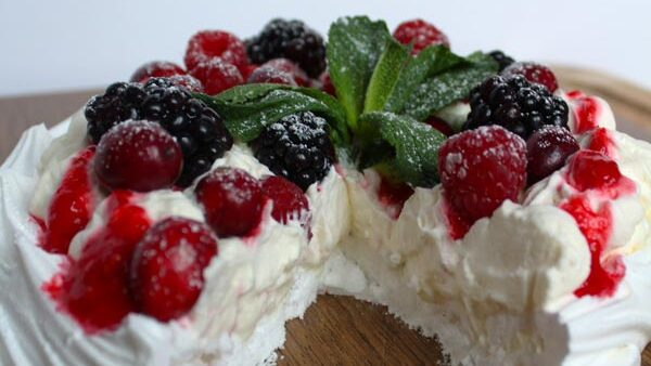 A pavlova has been cut and is missing a slice. It is topped with fresh raspberries and blackberries. A rich vegan whipped cream covers the outside of the pavlova and the inside contains a lush vegan meringue.
