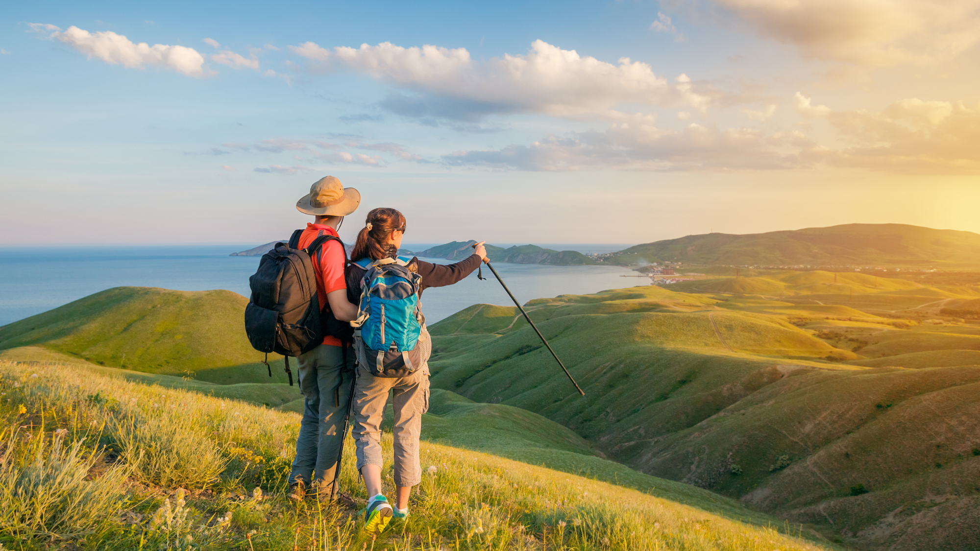 Two hikers with backpacks stand on a grassy hill at sunset, overlooking rolling green hills 