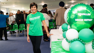 Pam is standing next to a table decorated for a Coffee Morning. She has short dark hair and is wearing dark trousers and a Macmillan branded green top. On the table are several green and white balloons. Behind Pam are different people attending the event.