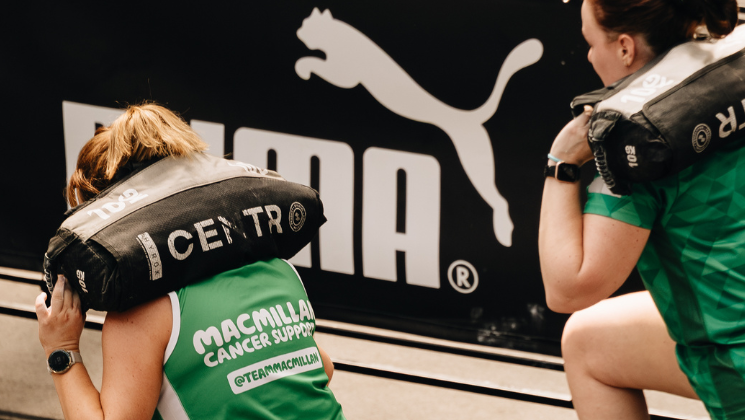 Two people in green Macmillan branded vest tops are doing lunges with sand bags on their backs.