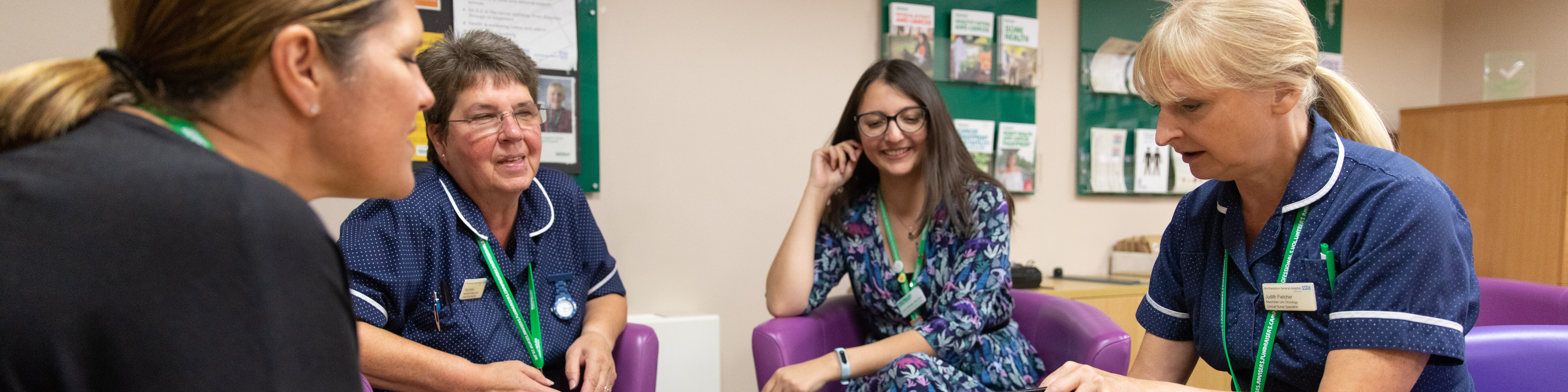 Four people sit around a small round table in a room, three in uniforms and one in casual clothing. One person uses a laptop while green Macmillan Cancer Support boards with posters and leaflets are visible in the background.