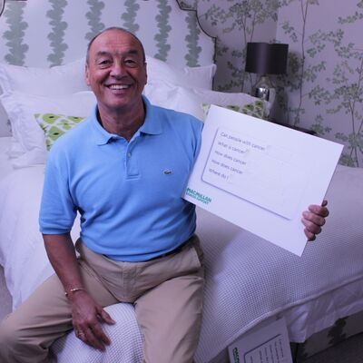 Elvin is sitting on a bed, looking at the camera and smiling. He is holiday a white board with questions about cancer written on it.