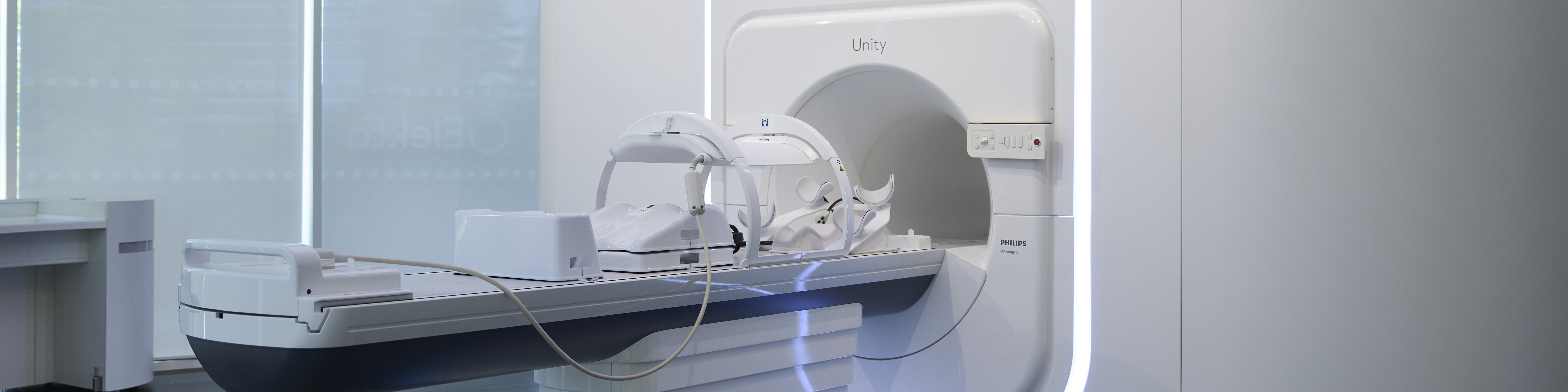 white Elekta MRI scanner with blue accent lighting stands in a bright, clean room with large windows and blinds. 