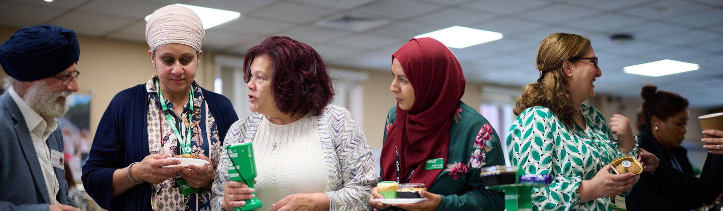 A group of people are standing and talking to one another at an event. In front of them is a table decorated for a Macmillan Coffee Morning. Some people are holding plates with food on them.