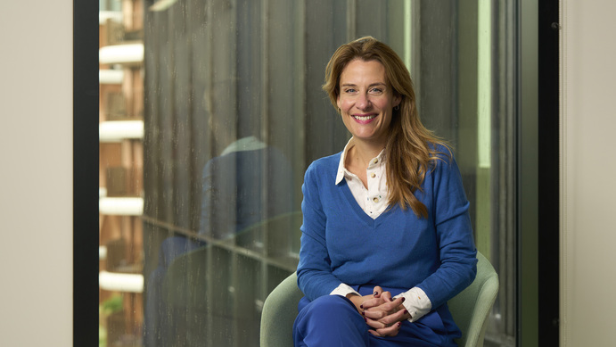 Gemma Peters sat on a chair in front of a window, smiling and wearing a white shirt, blue jumper and blue trousers