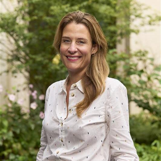 Gemma Peters is the CEO of Macmillan Cancer Support. She has long hair and is smiling. She is standing outside and has one hand placed on a rail. She is wearing a white shirt and blue trousers.