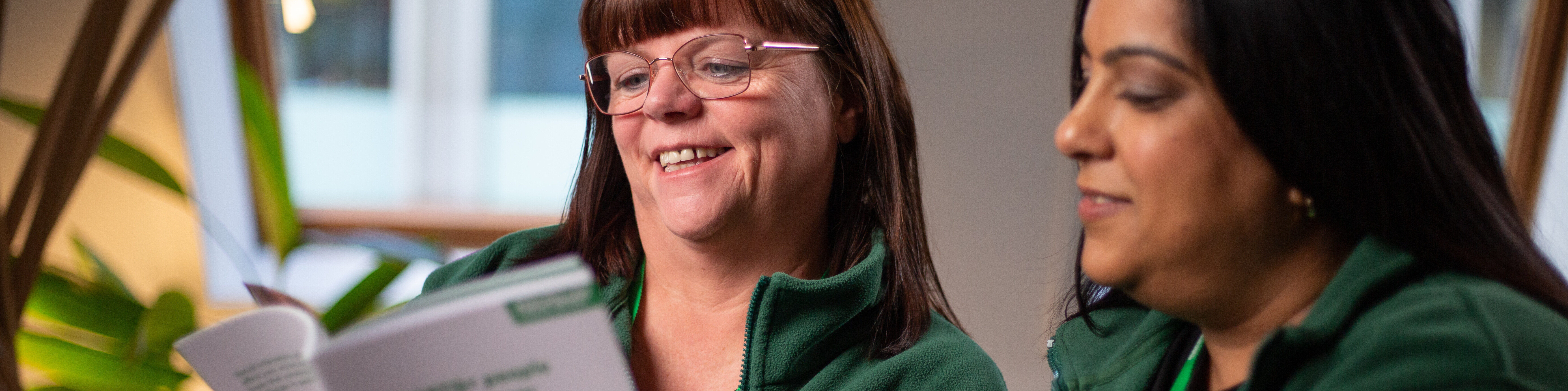 Two Macmillan employees reading a Macmillan booklet. They both are wearing green Macmillan tops and one is wearing glasses.