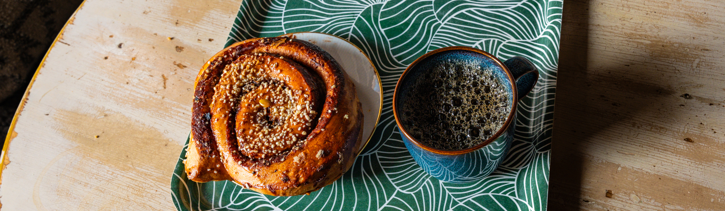 A colourful tray on a wooden table. Place on the tray is a large cinnamon bun on a plate and a dark cup of coffee.