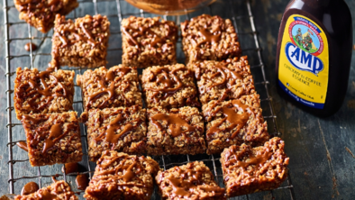 A batch of rich, golden-brown flapjacks cut into neat squares and arranged on a cooling rack. Each piece has a slightly crisp edge and a chewy oat texture, with a glossy drizzle of mocha icing zigzagged across the top. The icing, made with Camp Coffee, adds a deep coffee aroma and a hint of sweetness. These indulgent treats look homemade and comforting — perfect for coffee lovers seeking a twist on a classic bake.