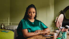 A young Black woman is sitting at home at a table which has make up on. There is a bed in the background with children's toys and books on.