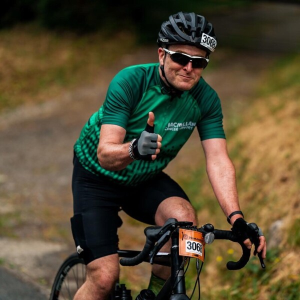 Man riding his bike with a Macmillan cycling jersey on and giving a thumbs up to the camera