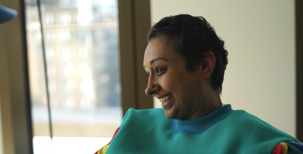 A woman with a colourful top, short hair and bright eye make-up is smiling