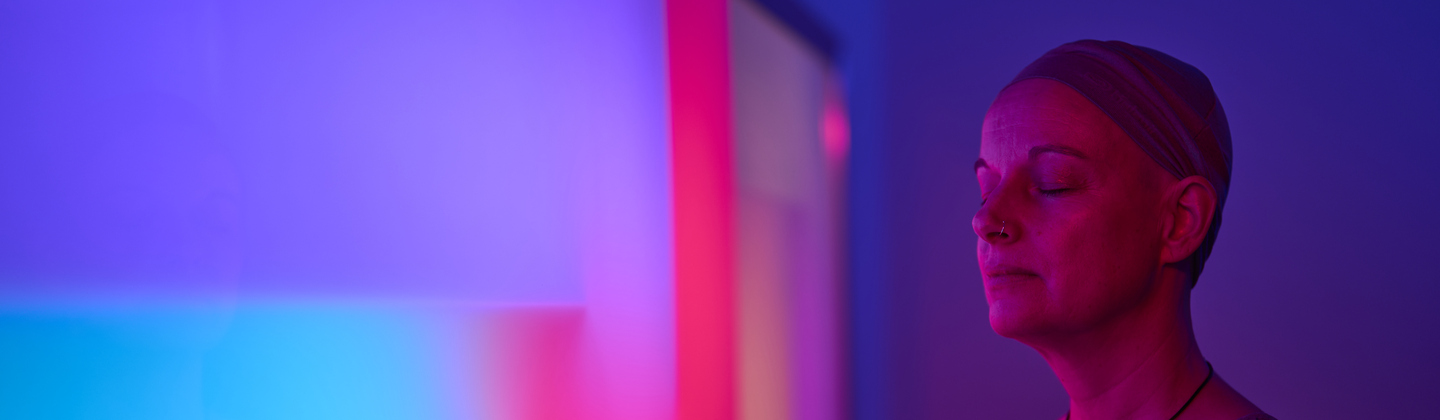 A women has her eyes closed and is relaxing whilst experiencing the Sanctuary Light and Sound Therapy room at the Macmillan Horizon Centre in Brighton. She has on a grey top and is wearing a head wrap. In front of her is a colourful wall on display.