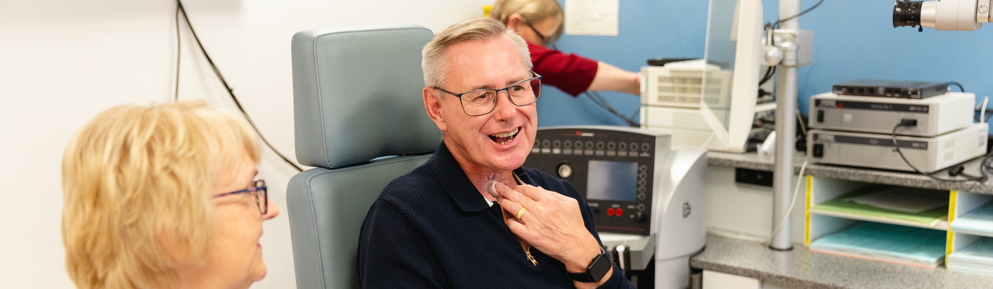 A person is sitting down on a chair in a clinic. They are speaking using a voice prostheses. Next to them is another person who is sitting down and listening to them speak. 