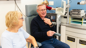 A person is sitting down on a chair in a clinic. They are speaking using a voice prostheses. Next to them is another person who is sitting down and listening to them speak. 