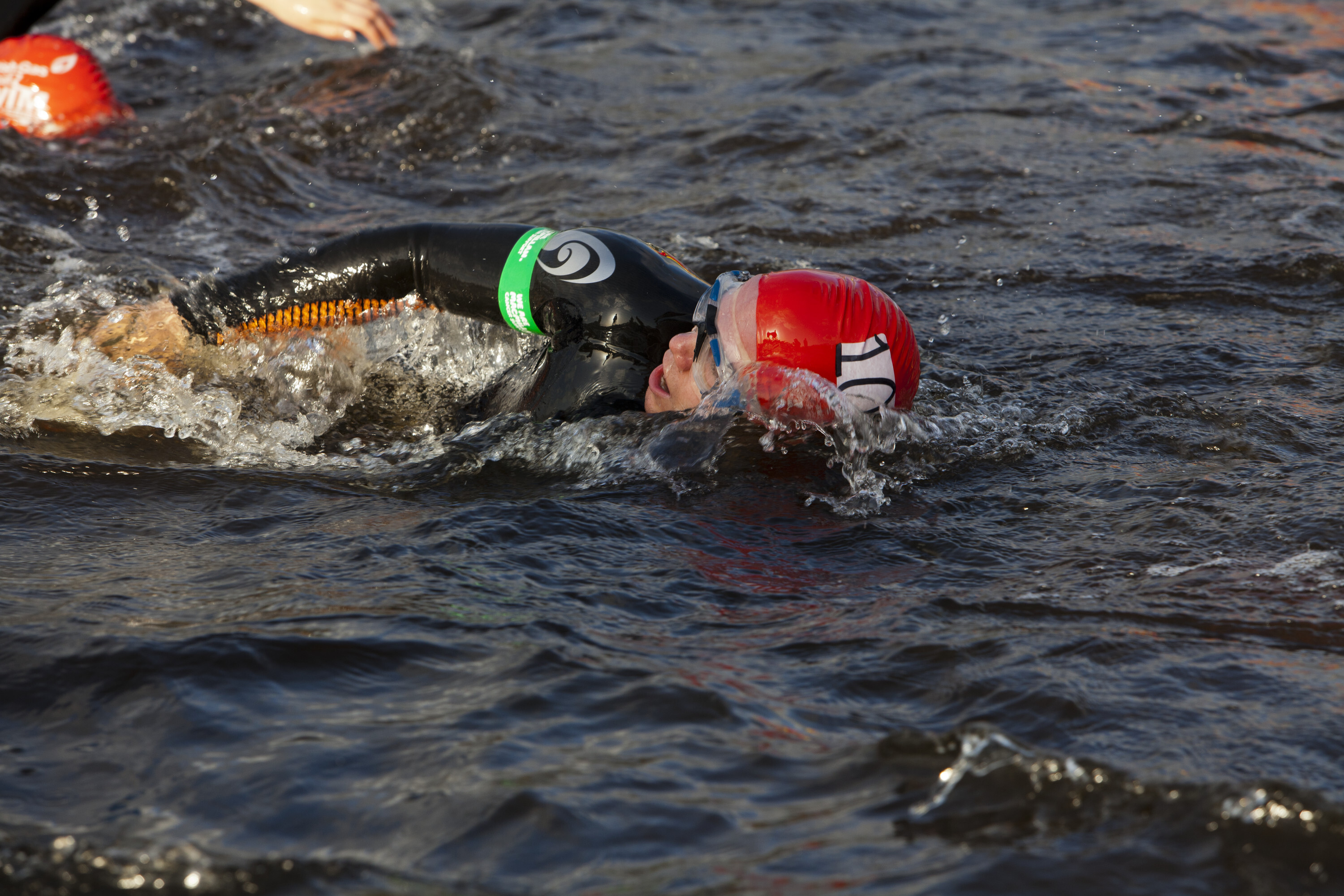 A person open water swimming. They wear a red swim cap and a green Macmillan armband.