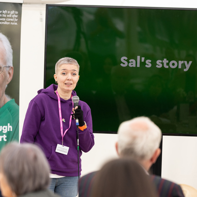 Sal is standing on a stage and is speaking into a microphone. She is wearing a dark purple hoodie and blue jeans. Behind Sal is a large screen with the words Sal's story on it. There is also a Macmillan branded banner. There is a crowd of people sitting and listening to Sal's story.