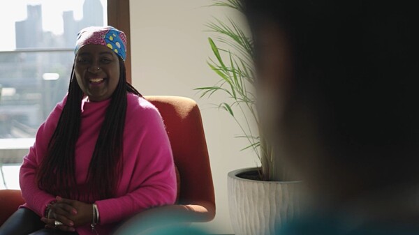 A Black woman in a bright outfit is smiling at someone. We can see the back of the other person's head.