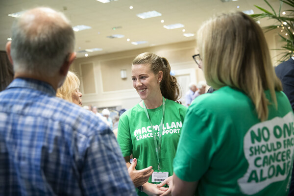 Group of 4 individuals stood in a circle talking. Woman in main focus is smiling wearing a green Macmillan tshirt and lanyard 