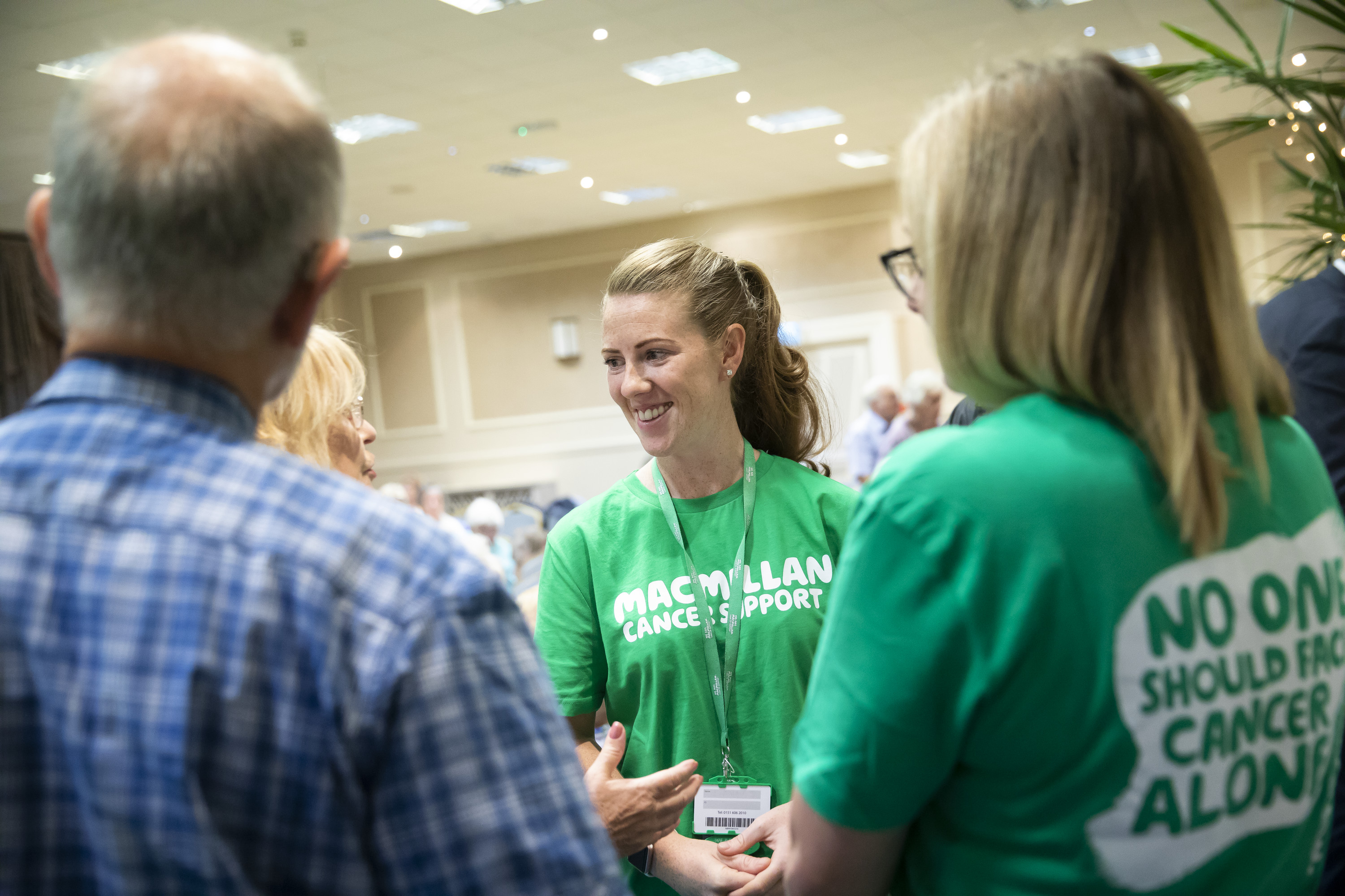 Group of 4 individuals stood in a circle talking. Woman in main focus is smiling wearing a green Macmillan tshirt and lanyard 