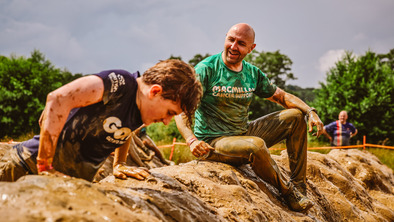 A smiling man wearing a Macmillan t-shirt sat on a mud bank supporting someone else trying to climb over it