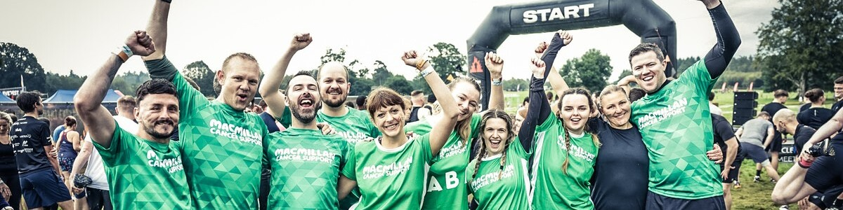 A group of 10 people at the start of their Tough Mudder with their hands in the air and wearing green Macmillan t-shirts. 