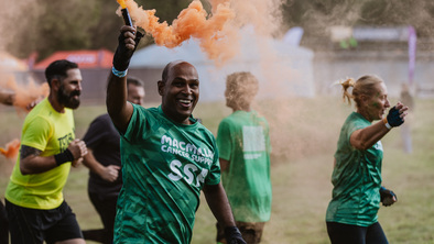 A man wearing a a green Macmillan t-shirt and letting off an orange smoke flare