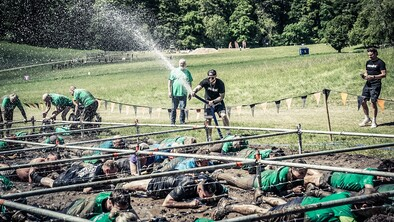 Someone spraying water over lots of people crawling under a barbed wire obstacle