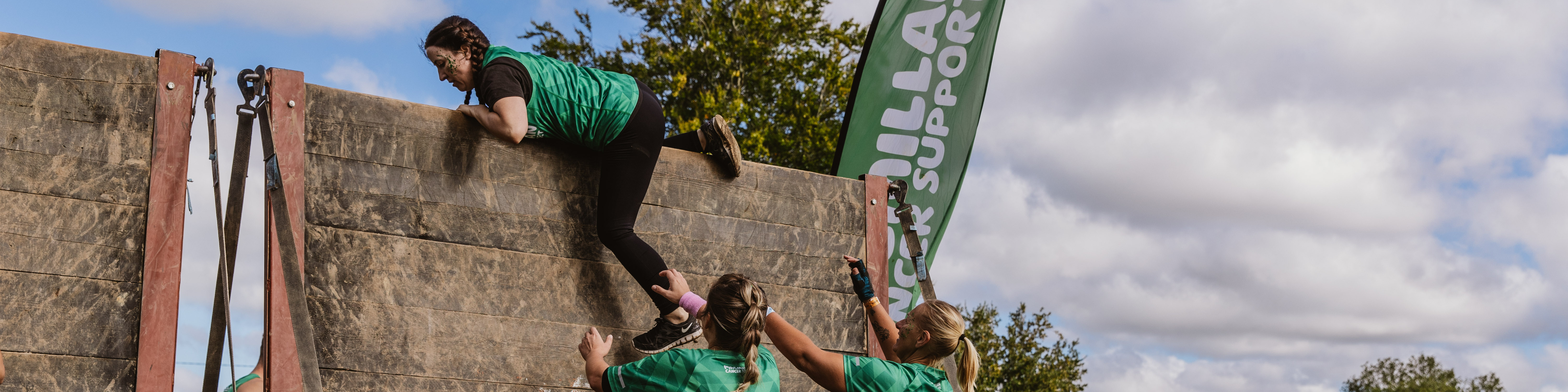 Two women wearing Macmillan t-shirts helping another over a wall obstacle 