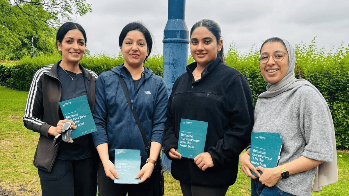 Group of four people standing outdoors on a paved path, each holding a teal booklet, with greenery and a blue lamp post in the background.