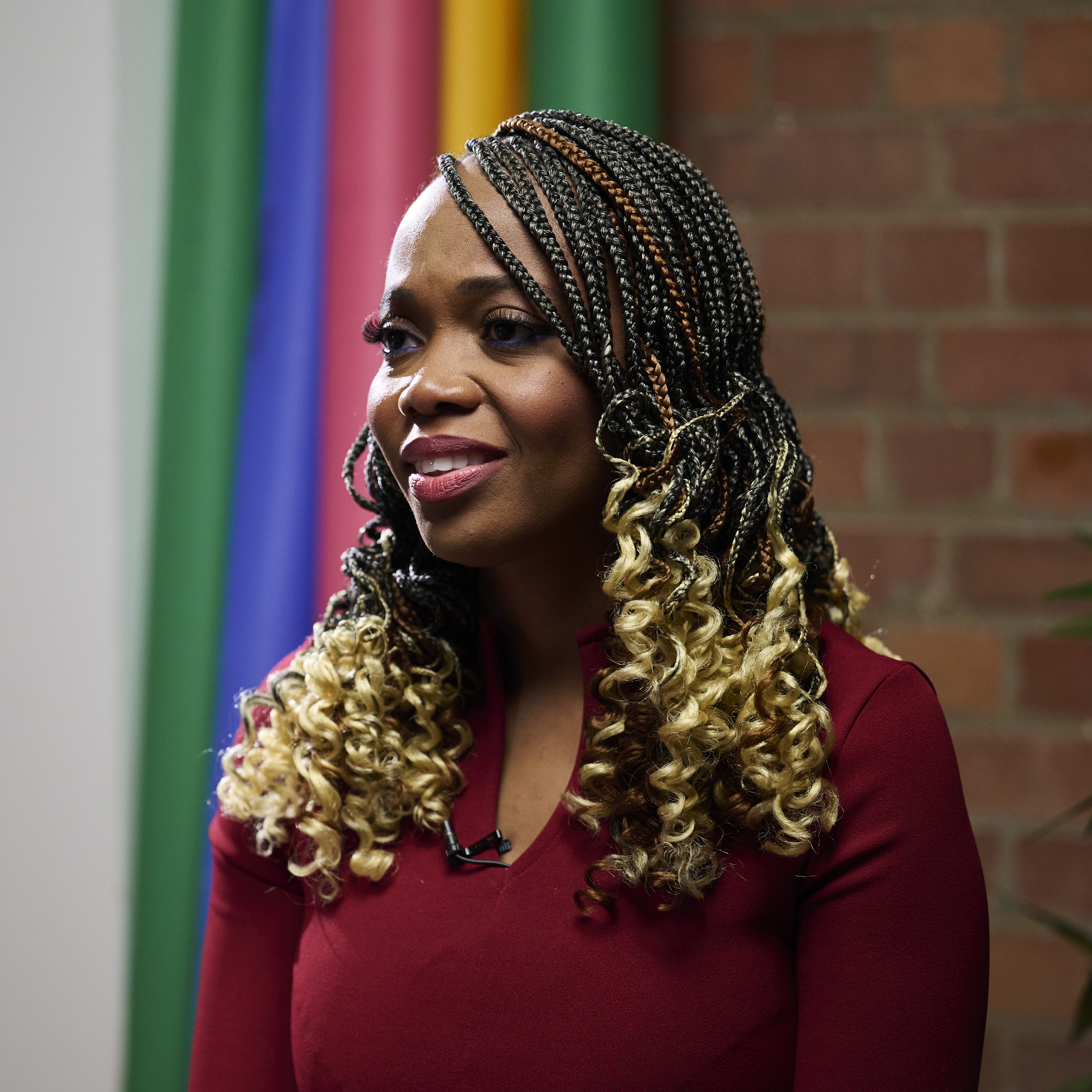 A person wearing a deep red long-sleeved top sits indoors in front of a backdrop of vertical rainbow-coloured stripes. Their hair is styled in long, dark braids with golden curls at the ends. A brick wall and green plant are visible in the background, creating a modern, vibrant setting.
