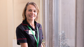 Claire is wearing a dark uniform with red piping and a green lanyard. She is standing indoors near a large window, resting both hands on a wooden railing. 