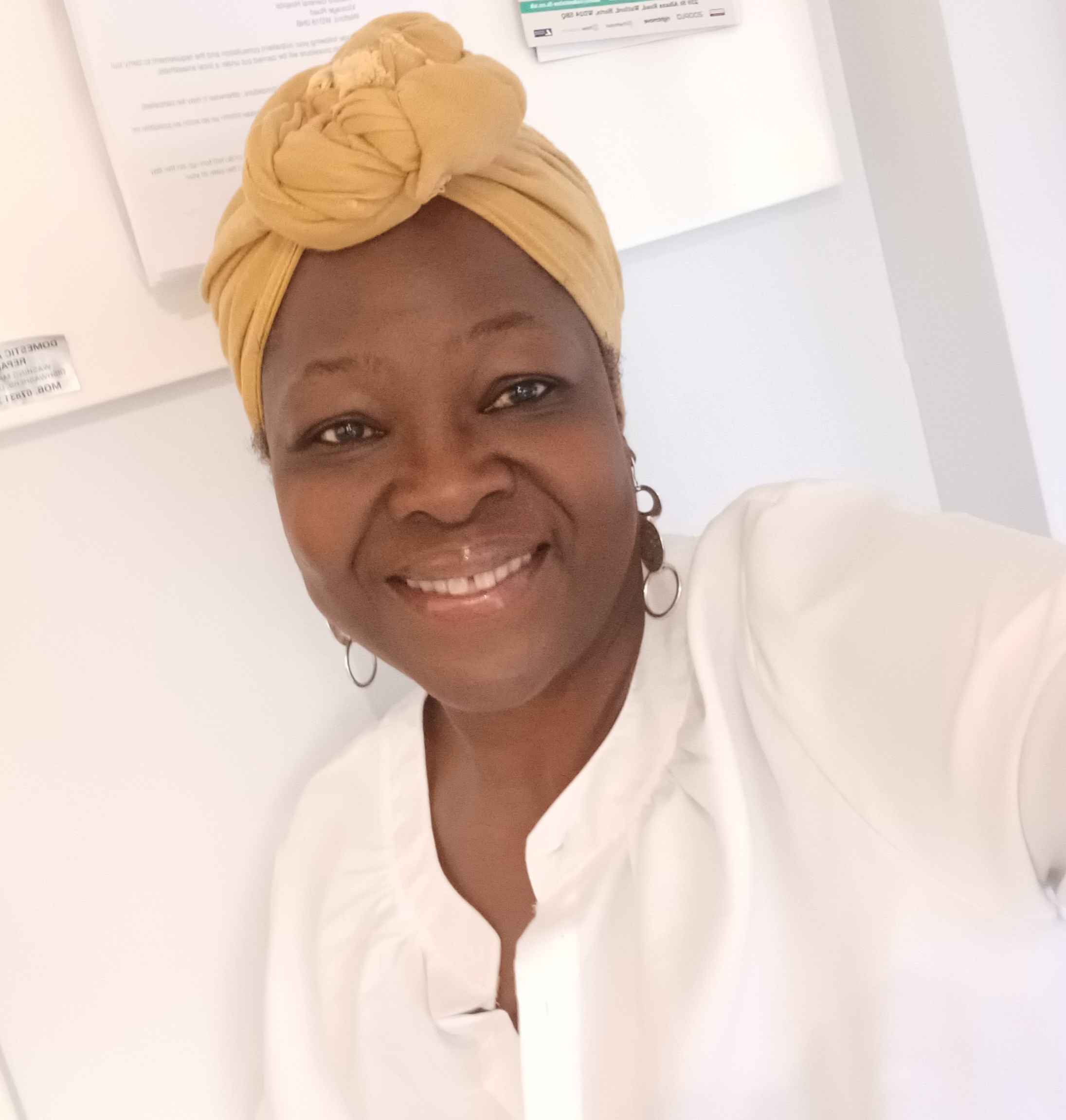 Woman smiles at camera, she is wearing a white shirt and mustard coloured headwrap