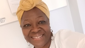 Woman smiles at camera, she is wearing a white shirt and mustard coloured headwrap