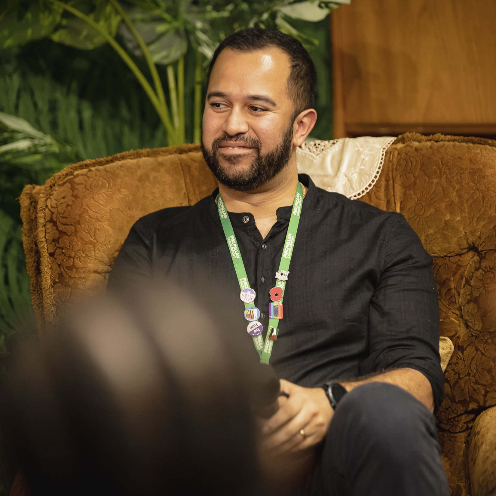 Speaker seated on a vintage armchair at Macmillan Cancer Support Open House, wearing event lanyard.
