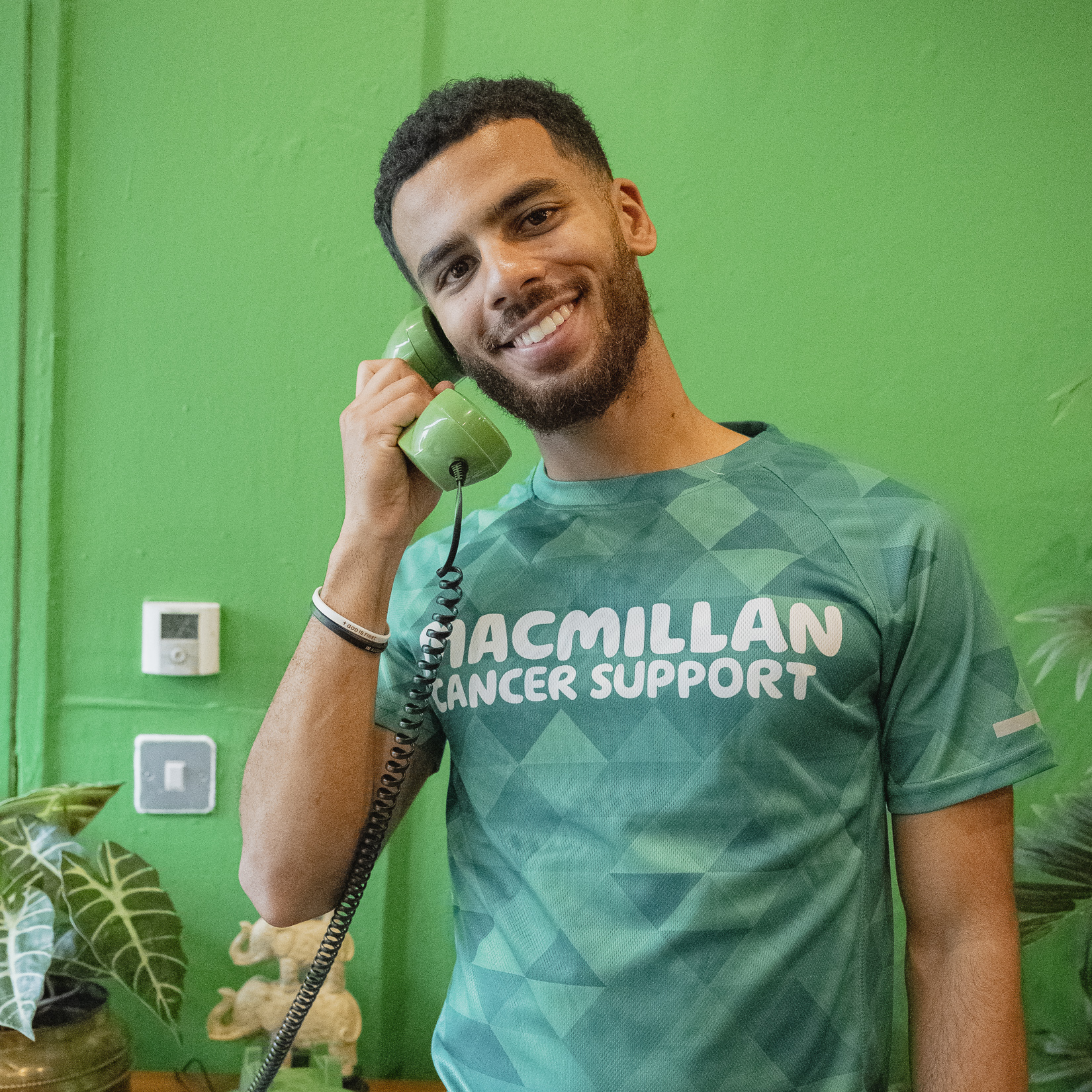 Person wearing a green Macmillan Cancer Support T-shirt, holding a green retro-style telephone receiver to their ear. They are standing indoors against a bright green wall, with a wooden sideboard in front that has a matching green rotary phone and potted plants.
