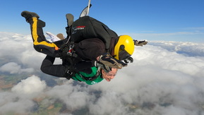 Two people strapped together skydiving above clouds with blue sky in the background