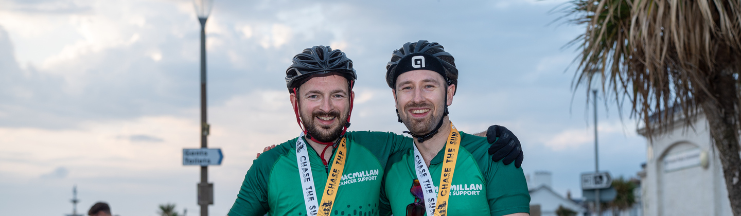 Two people are standing side by side. They both are wearing green Macmillan branded cycling tops, bicycle helmets and other cycling gear on. They also have Chase the Sun finishers medals around their necks, and are holding up a bicycle with one arm. They appear to be outside and at the end of the Chase the Sun event.