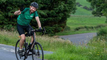 A cyclist is smiling and waving whilst riding their bicycle. They are on a paved road through the countryside. They are wearing cycling gear, including a green Macmillan branded cycling top.