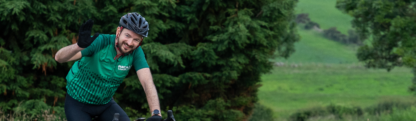 A cyclist is smiling and waving whilst riding their bicycle. They are on a paved road through the countryside. They are wearing cycling gear, including a green Macmillan branded cycling top.