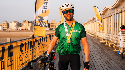A cyclist is standing on a beach pier propping up a bicycle with their right arm. They are wearing cycling gear including a green Macmillan branded cycling top. They also have a Chase the Sun finishers medal around their neck. 